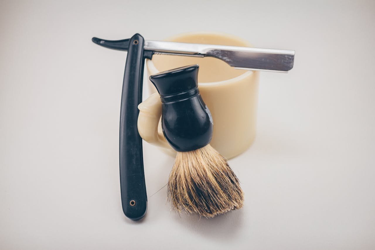 experience-img Still life of barber tools including a straight razor, shaving brush, and mug.